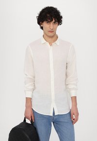 Bela lanena srajca z dolgimi rokavi, v kombinaciji z modrimi jeans hlačami. Model drži črn teksturiran nahrbtnik. Minimalistična zasnova, lahka tekstura.