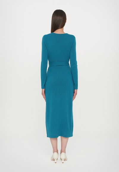Teal långärmad klänning med figursydd midja och rak silhuett, gjord av strukturerat tyg, kombinerad med cremefärgade geometriska klackar.