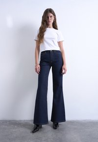 Γυναίκα με μακριά μαλλιά στέκεται μπροστά σε λευκό τοίχο φορώντας λευκό κοντομάνικο τοπ, σκούρο τζιν ψηλόμεσο με άνοιγμα, μαύρα μυτερά μποτάκια και ασημένιο βραχιόλι.