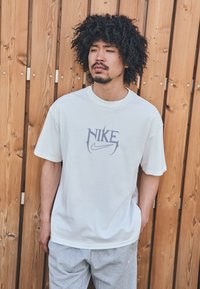 Hvid bomulds T-shirt med et lilla Nike-logo, der har korte ærmer og en klassisk pasform, parret med grå sweatpants. Træbaggrund.