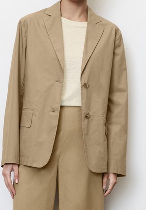 Personne portant une veste de costume beige à deux boutons sur un haut crème clair et un pantalon beige assorti, debout devant un fond uni.