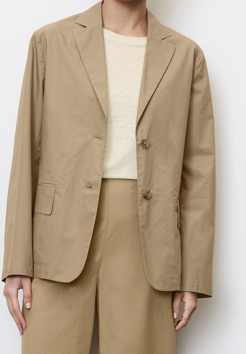 Personne portant une veste de costume beige à deux boutons sur un haut crème clair et un pantalon beige assorti, debout devant un fond uni.