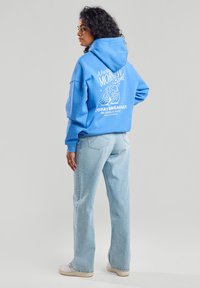 Light blue hoodie with white graphic print on the back, featuring bagel design and text. Paired with light blue straight-leg jeans and sneakers.