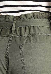 Olivgröna byxor med en samlad midja, sidoficka och subtil textur, som är kombinerade med en svart och vit randig topp.