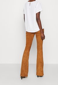 Blouse blanche à manches courtes avec une ouverture en forme de goutte dans le dos, associée à un pantalon évasé marron en suédine, présentant une texture lisse et une coupe ajustée.