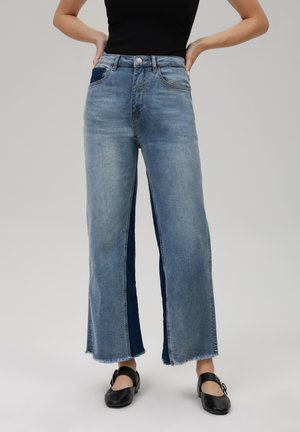 Jeans de cintura alta y pierna ancha en denim azul claro deslavado con paneles laterales en azul oscuro y dobladillos sin acabar. Incluye bolsillos frontales. Se llevan zapatos negros.