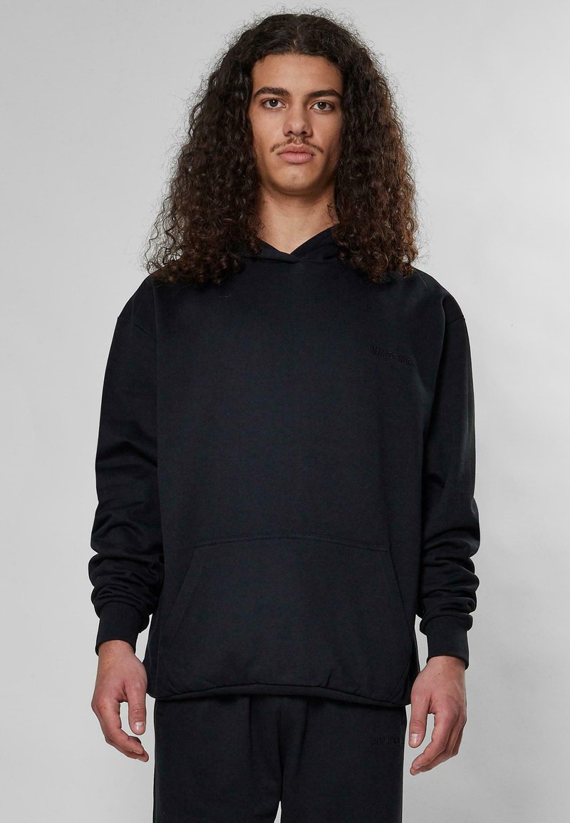 Sudadera negra de tejido suave, con un bolsillo frontal, puños acanalados y capucha con cordón. Diseño minimalista sin logotipos visibles.