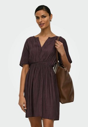 Ženska v temno vijoličasti teksturirani obleki, dolgi do kolen s kratkimi rokavi, nosi veliko rjavo usnjeno torbo čez ramo.