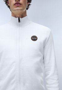 Vit zip-up tröja med texturerad kropp, hög krage och en cirkulär logotyp-patch med orange och svarta detaljer på bröstet.