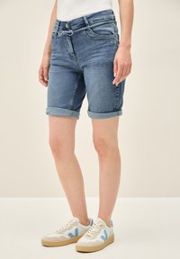 Shorts de mezclilla en azul claro con dobladillo enrollado, con bolsillos delanteros y cintura con cordón. Combinados con zapatillas blancas con detalles en azul.