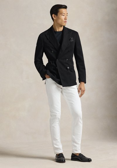 Άνδρας που φοράει μαύρο διπλό σακάκι, μαύρο πουκάμισο, λευκό παντελόνι και μαύρα μοκασίνια, στέκεται μπροστά σε απλό ουδέτερο φόντο.