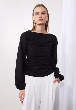 Black long-sleeve blouse with a gathered texture and boat neckline, paired with white wide-leg trousers. Model wears statement earrings.