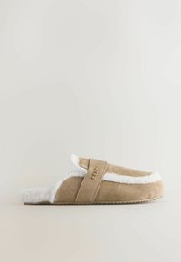 Tan suède slipper met witte nepbont voering, open achterkant en verstelbare band. Beschikt over een gestructureerde zool en een metallic logo accent.