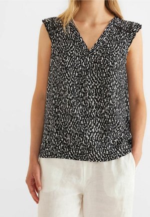 Vrouw die een mouwloze zwarte en witte blouse met V-hals en patroon draagt, gecombineerd met witte broek, staand met één hand in de zak.