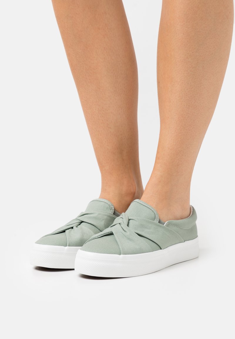Zapatos slip-on de lona verde con detalles anudados en la parte superior, que cuentan con una suela de goma blanca texturizada para mayor agarre.