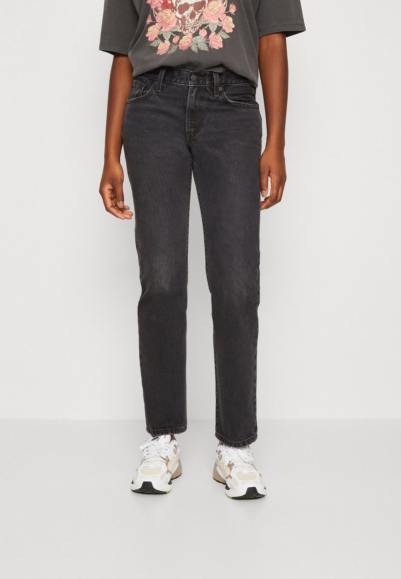Persona que lleva unos vaqueros negros desgastados de pierna recta, zapatillas blancas y una camiseta oscura con un diseño de cráneo y flores rosas.