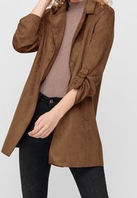 Trench-coat couleur sable, aspect daim, avec col à revers, manches retroussées et coupe décontractée, associé à un haut marron côtelé et un jean noir.