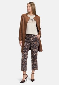Cappotto in suede sintetico marrone, t-shirt grafica beige con dettagli metallici, pantaloni cropped a motivi marroni e blu, e tacchi alti scuri con cinturini alla caviglia.