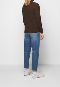 Brązowy sweter z dzianiny z wysokim kołnierzem, w połączeniu z jasnoniebieskimi spodniami o prostym kroju i beżowymi trampkami. Tekstury są miękkie i casualowe.