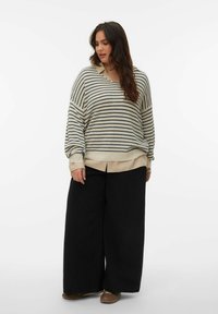 Femme aux longs cheveux noirs portant un pull beige rayé, une chemise beige, un pantalon large noir et des chaussures marron, debout sur un fond blanc uni.