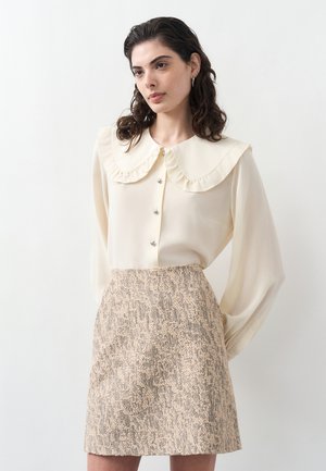 Frau trägt eine cremefarbene Bluse mit großem, gerafftem Kragen und einen strukturierten beigefarbenen A-Linien-Rock vor einem schlichten weißen Hintergrund.