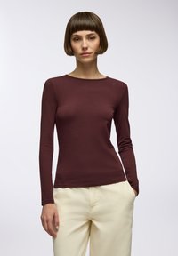 Burgundowa bluzka z długim rękawem w dopasowanym kroju z okrągłym dekoltem, wykonana z gładkiego, lekkiego materiału; zestawiona z jasnymi spodniami.