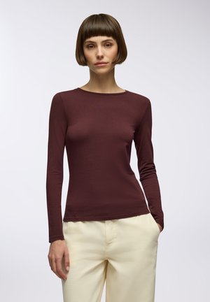 Maglietta bordeaux a maniche lunghe dal design aderente con scollatura rotonda, realizzata in un tessuto liscio e leggero; abbinata a pantaloni chiari.
