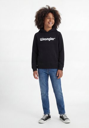 Mosolygós gyerek göndör hajjal, fekete Wrangler kapucnis pulóvert, kék farmert és fekete magas szárú tornacipőt visel, fehér háttér előtt áll.