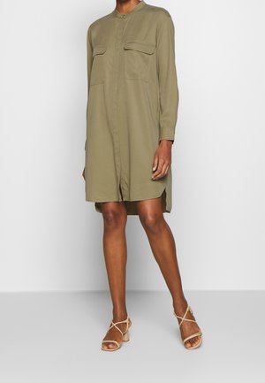 Kobieta ubrana w luźną sukienkę koszulową z długim rękawem w kolorze oliwkowej zieleni, z klapkowymi kieszeniami z przodu i beżowymi sandałami na niskim obcasie z paskami.