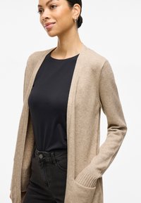 Beige strikkejakke med åpen front, ribbestrikkede mansjetter og sidelommer, brukt over en mørk topp og slim fit svarte bukser. Glatt tekstur.