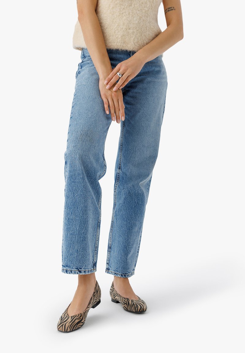 Personne portant un jean droit bleu clair, un haut sans manches beige duveteux, des ballerines à imprimé zèbre et une bague en argent, se tenant les bras croisés.