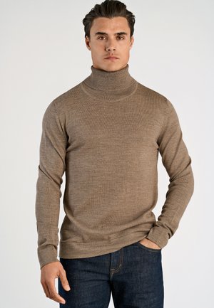 Beżowy sweter z golfem o miękkiej, prążkowanej fakturze, z długimi rękawami, dopasowanym krojem i subtelnym logo na piersi. Noszony z ciemnymi dżinsami.