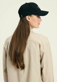 Czarna czapka baseballowa z wygiętym daszkiem i regulowanym paskiem. Noszona z jasnobeżową koszulą z guzikami, ukazującą długie, proste włosy.