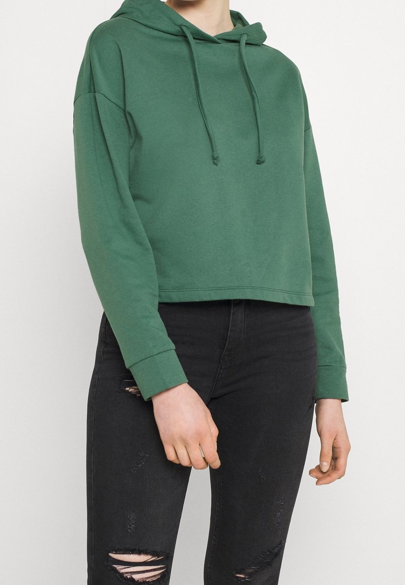 Osoba v zelené zkrácené mikině s kapucí a černých roztrhaných skinny džínech, stojící před bílým pozadím.