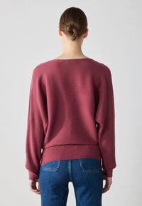 Burgunderfarget ribbestrikket genser med lange ermer og en avslappet passform; har en vid utrigning og en kort hem. Kombinert med blå jeans.
