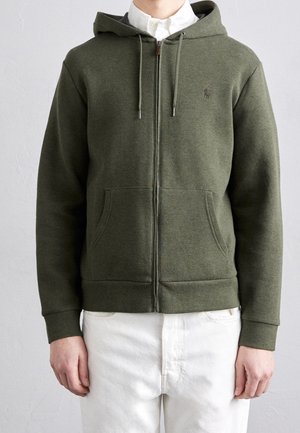 Henkilö, joka pukeutunut oliivinvihreään vetoketjulliseen hupulliseen huppariin, jossa on etutaskut ja kiristysnauhat, yhdistettynä valkoisiin housuihin ja valkoiseen napilliseen paitaan.