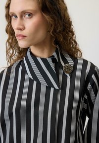 Ženska s kodrastimi lasmi, oblečena v črno-belo srajco z navpičnimi črtami in edinstvenim ovratnikom ter okroglem kovinskim gumbom blizu vratu.