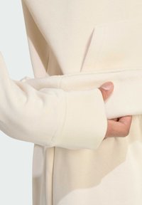 Felpa beige chiaro con una morbida texture, dotata di tasca a marsupio e polsini a coste. Primezza di una mano che afferra l'orlo.