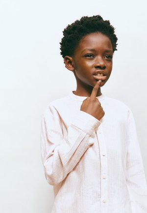 Persona giovane con capelli ricci che indossa una camicia chiara abbottonata, toccandosi le labbra con un dito, con uno sguardo pensieroso su uno sfondo semplice.