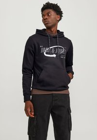Sudadera negra con bolsillo frontal, que presenta un logotipo y diseño gráfico en blanco de "Jack & Jones". Se lleva con pantalones cargo oscuros a juego.