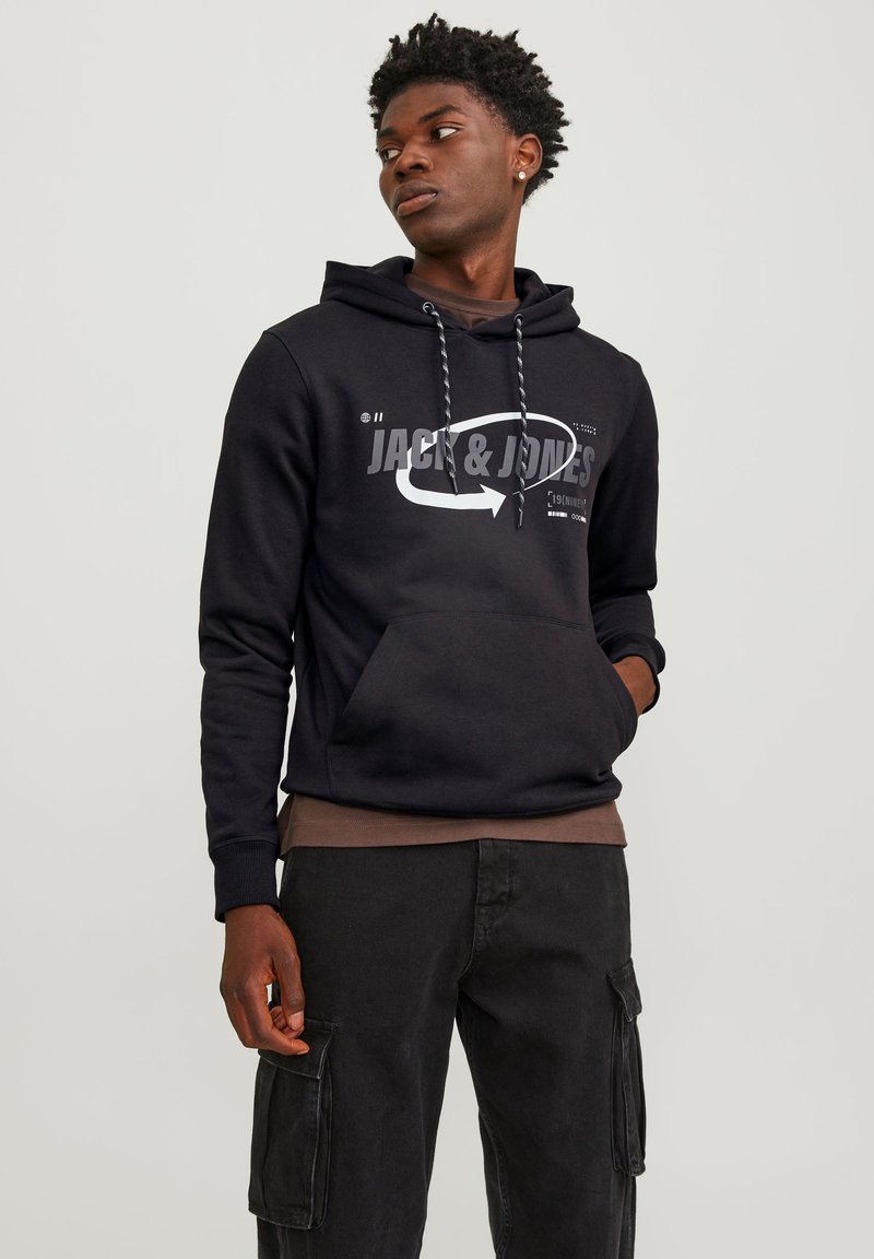 Sudadera negra con bolsillo frontal, que presenta un logotipo y diseño gráfico en blanco de "Jack & Jones". Se lleva con pantalones cargo oscuros a juego.