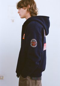 Tmavomodrá mikina s kapucňou a logami Toyota. Má klokanie vrecko a rebrované manžety. Nosená cez kockovanú košeľu a béžové nohavice.