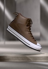 Brown leather high-top sneaker with white rubber sole, lace-up front, and contrasting toe cap. Minimalist design with subtle stitching details.