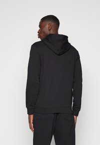 Svart hoodie med en stor huva och sidofickor, med en avslappnad passform. Tyget verkar slätt, med synliga stygn som detaljer vid sömmarna.