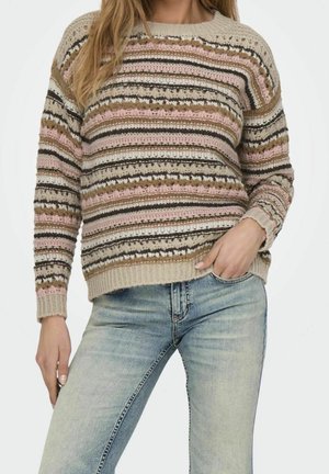 Femme portant un pull en tricot rayé multicolore et un jean bleu clair délavé, une main dans la poche, sur un fond uni.