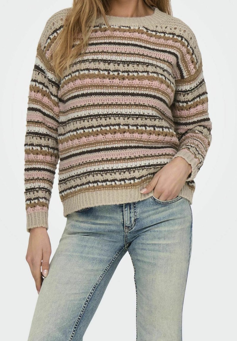 Femme portant un pull en tricot rayé multicolore et un jean bleu clair délavé, une main dans la poche, sur un fond uni.