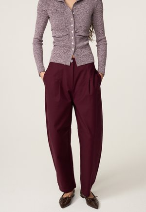 Vrouw draagt een aangesloten, lange mouwen, knoopsluitend topje van gestructureerde stof en een bordeauxrode, wijde, hoog getailleerde broek met zakken.