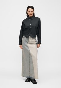 Γυναίκα που στέκεται φορώντας μαύρο πουκάμισο με κουμπιά, μακρύ τζιν φούστα με δύο αποχρώσεις και μαύρα παπούτσια σε απλό λευκό φόντο.