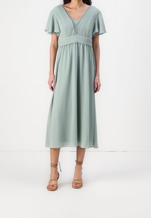 Kobieta ubrana w jasnozieloną sukienkę midi z plisowanym dekoltem w serek, falbanowymi rękawami, marszczoną talią i beżowymi sandałami na obcasie z paskami.