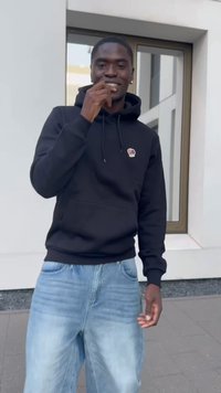 Black hooded sweatshirt with a small embroidered logo, paired with light blue baggy jeans. Standing outdoors with a neutral background.
